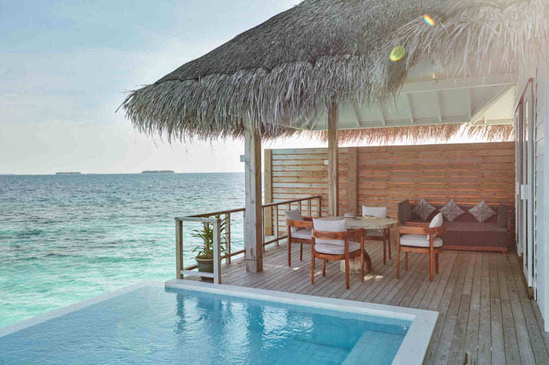 An overwater villa deck at Sun Siyam Iru Veli under a thatched roof, dining table and sofa beside the private pool.