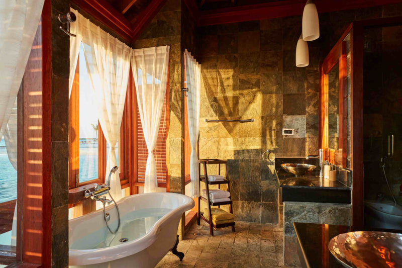 A stone and timber bathroom at Sun Siyam Olhuveli with a freestanding bath beside the lagoon windows.