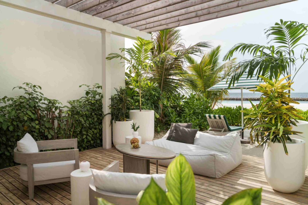 Sun Siyam Iru Veli, Dhaalu Atoll — shaded deck under a timber pergola with a low sofa and armchairs, potted palms, and a striped parasol on the sand beyond