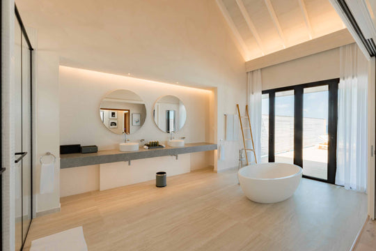 A villa bathroom at ananea Madivaru Maldives, twin round mirrors over a long vanity with a freestanding bath beside glazed doors