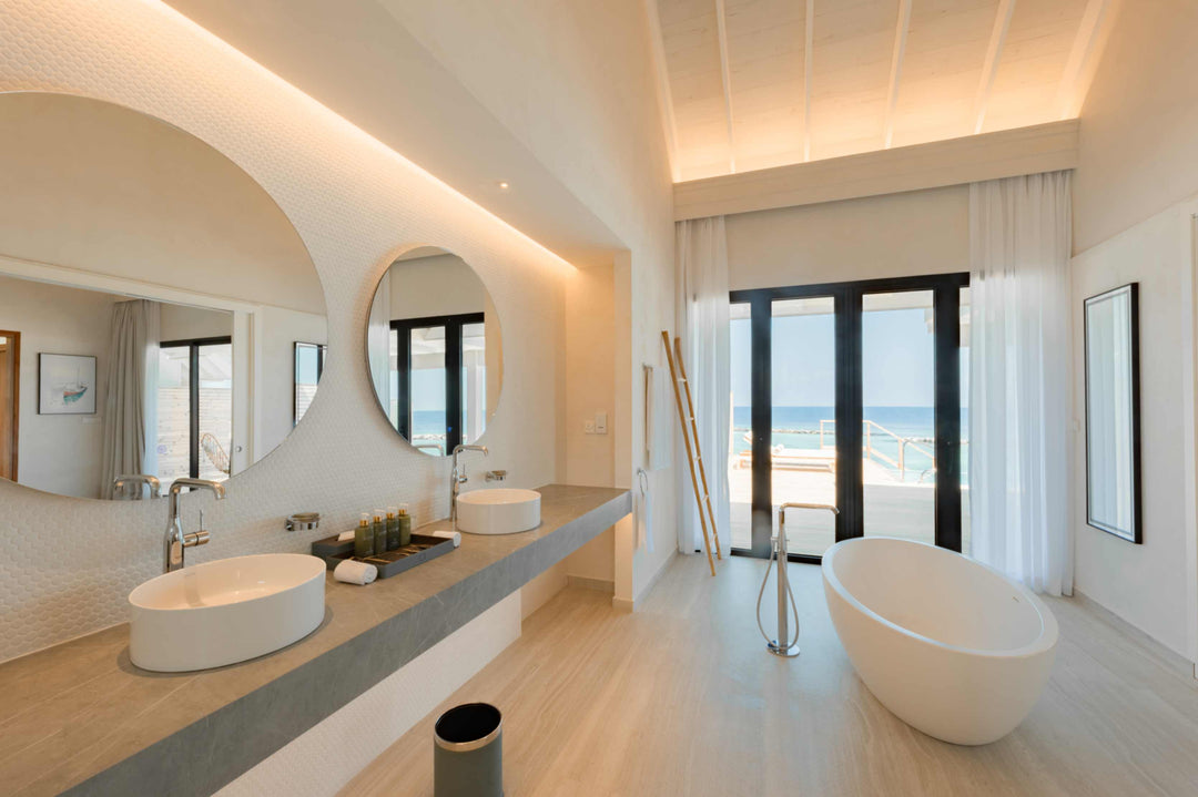 Twin basins under round mirrors in a villa bathroom at ananea Madivaru Maldives, a freestanding bath and the sea through the doors