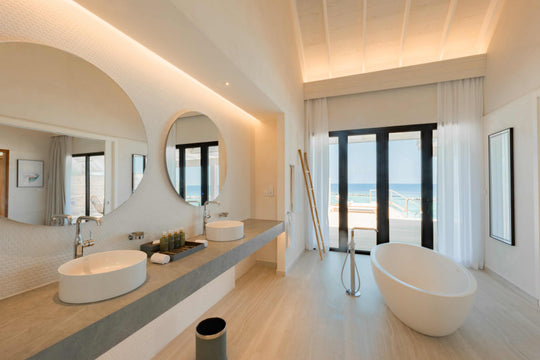 Twin basins under round mirrors in a villa bathroom at ananea Madivaru Maldives, a freestanding bath and the sea through the doors