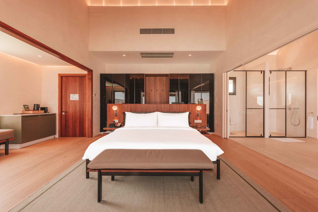 A villa bedroom at ananea Madivaru Maldives, the bed against a timber headboard wall between two glazed shower screens
