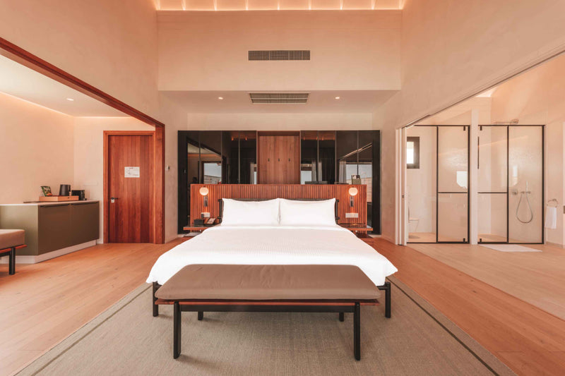 A villa bedroom at ananea Madivaru Maldives, the bed against a timber headboard wall between two glazed shower screens