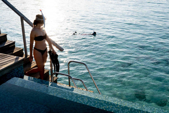 A snorkeller carrying fins steps down a ladder into the lagoon from an over-water deck
