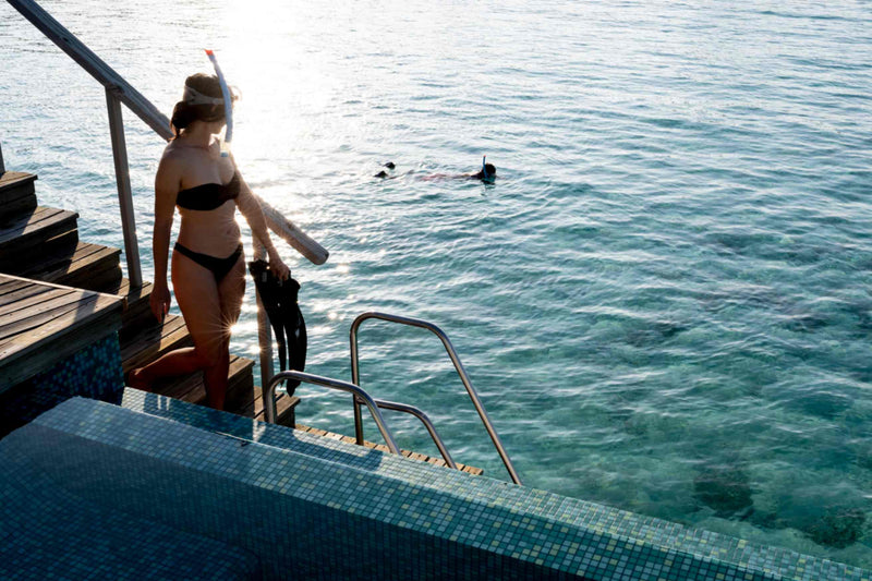 A snorkeller carrying fins steps down a ladder into the lagoon from an over-water deck at OUTRIGGER Maldives Maafushivaru Resort.