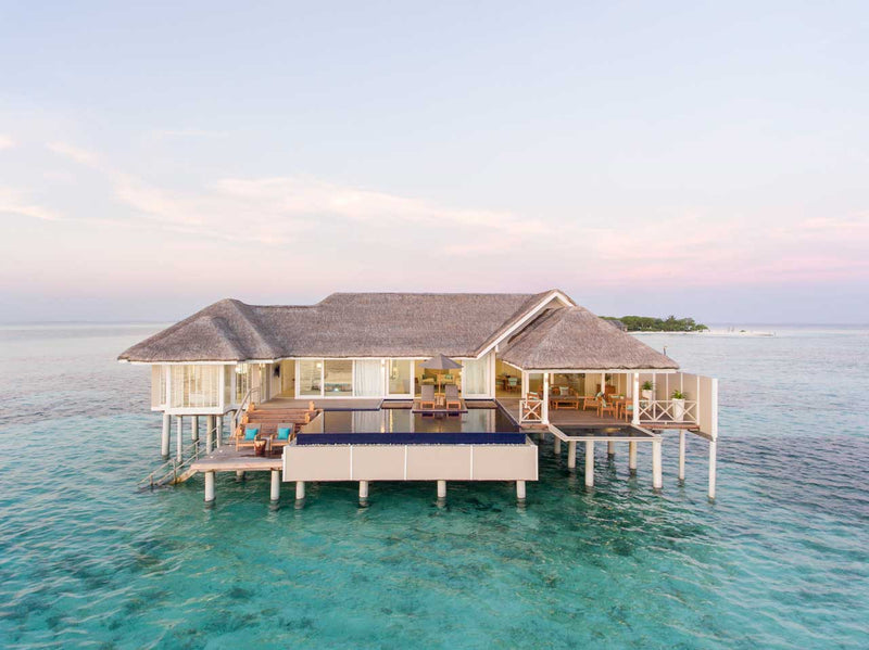A large villa at LUX* South Ari Atoll on stilts at dusk, pitched roofs and a pool on its deck.