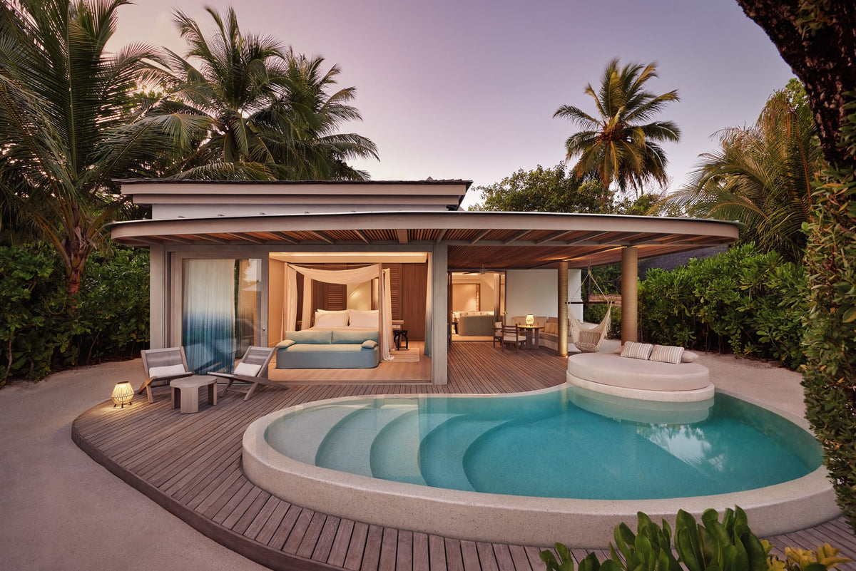 A beach villa at LUX* South Ari Atoll lit at dusk, curved pool on a timber deck among palms.