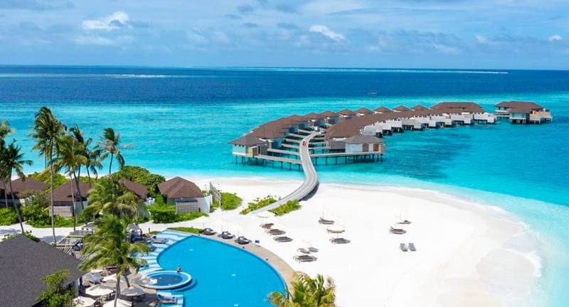 The main pool at Avani+ Fares Maldives, by the beach with palms and the overwater villa jetty beyond.