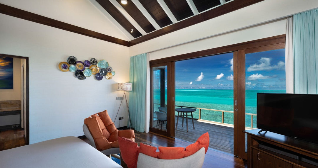 Atmosphere Kanifushi, Lhaviyani Atoll — inside an overwater villa: a bed and a sofa with orange cushions facing glass doors that open on to the deck and the turquoise lagoon.