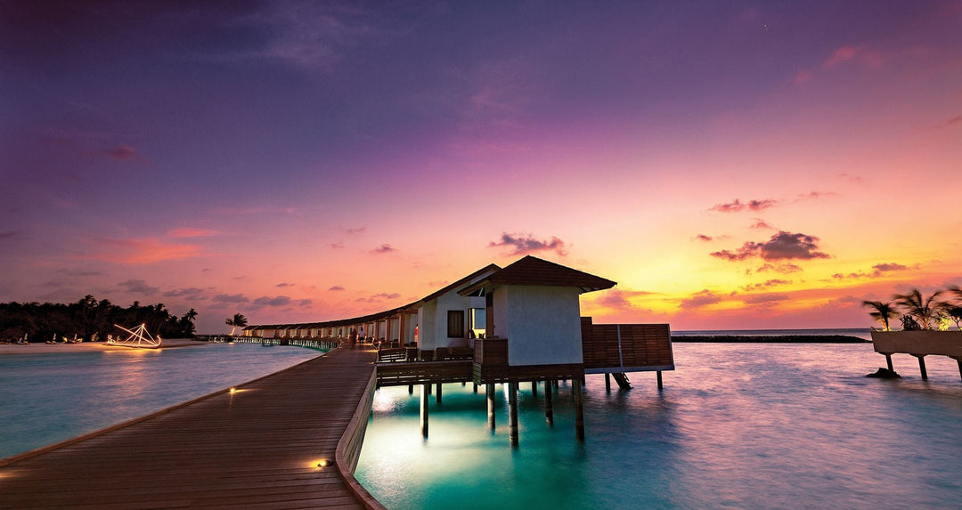 Atmosphere Kanifushi, Lhaviyani Atoll — the water villa jetty at sunset, a lit villa at the end of the boardwalk and more of them along the reef under a purple and orange sky.