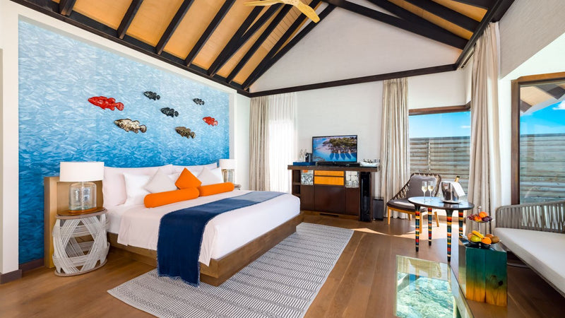 The bedroom of a Wind Villa at OZEN LIFE Maadhoo, a painted shoal of fish on the wall behind the bed and a glass floor panel beyond