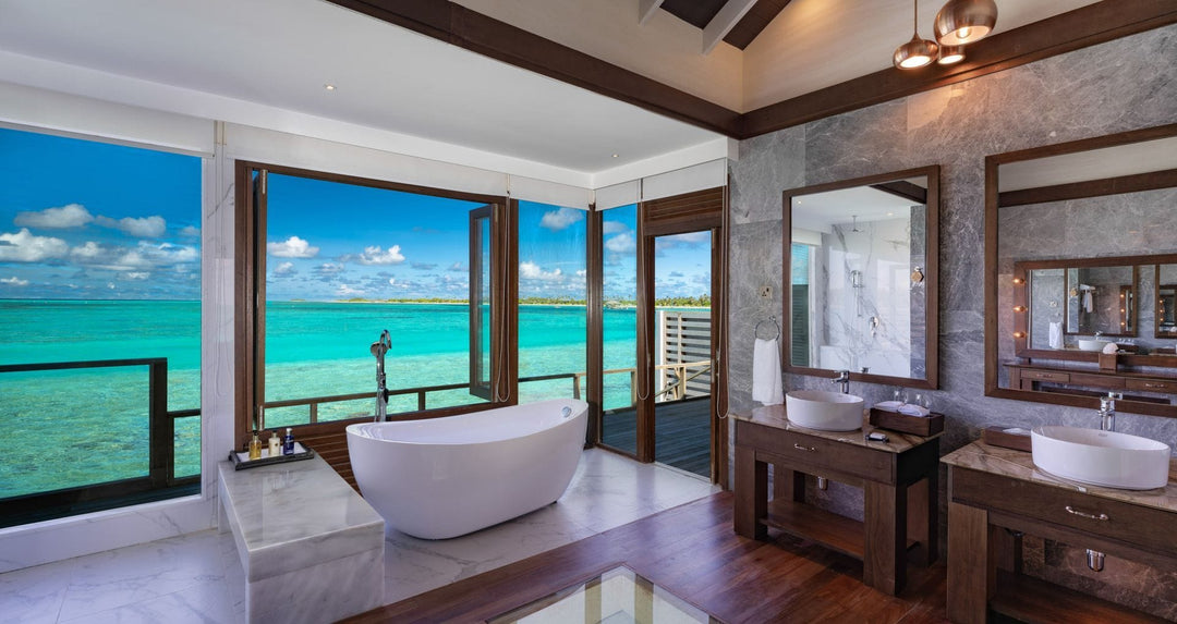 Atmosphere Kanifushi, Lhaviyani Atoll — an overwater villa bathroom: a freestanding bath at open windows over the lagoon, with twin stone basins and marble walls alongside.