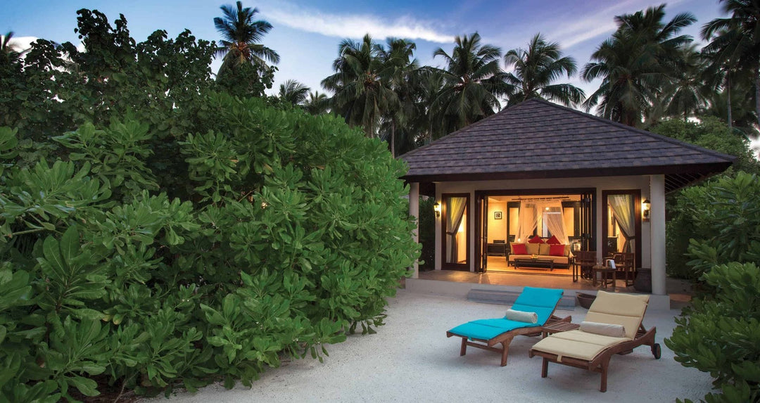 Atmosphere Kanifushi, Lhaviyani Atoll — a beach villa at dusk, lit from inside behind dense green vegetation, with two sun loungers set out on the sand in front of it.