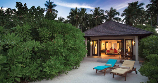 Atmosphere Kanifushi, Lhaviyani Atoll — a beach villa at dusk, lit from inside behind dense green vegetation, with two sun loungers set out on the sand in front of it.