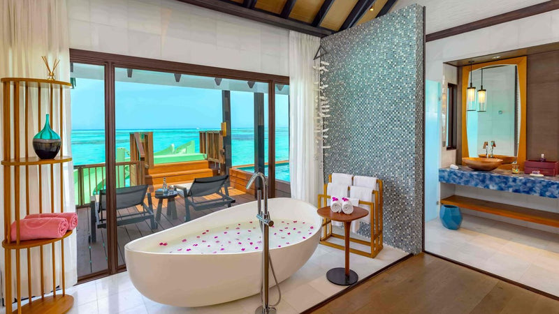 Inside a Wind Villa with Jacuzzi & Slide at OZEN LIFE Maadhoo, a freestanding bath scattered with petals and glass doors onto the deck