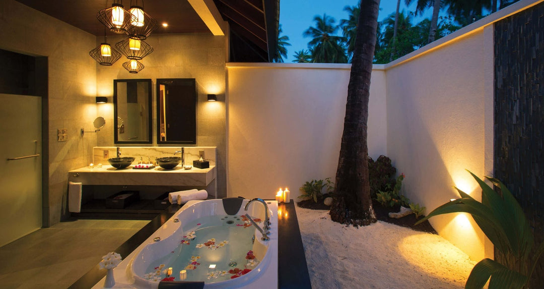 Atmosphere Kanifushi, Lhaviyani Atoll — a walled outdoor bathroom lit at night, a sunken bath scattered with petals and twin basins, with a palm growing up through the sand.