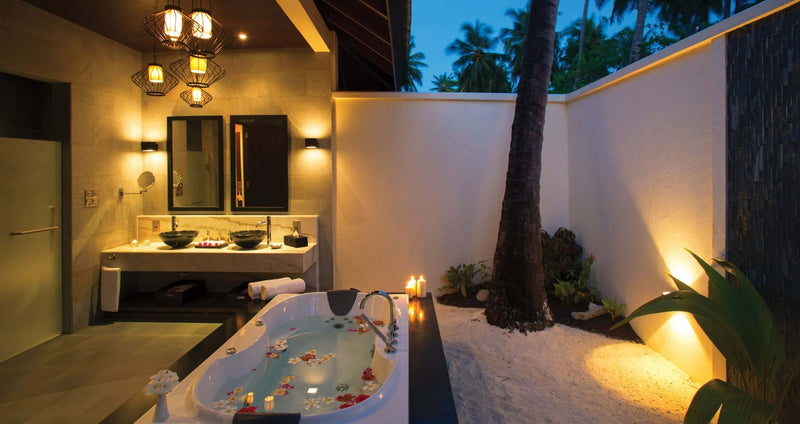 A walled outdoor bathroom at Atmosphere Kanifushi lit at night, a sunken bath scattered with petals and twin basins, with a palm growing up through the sand.