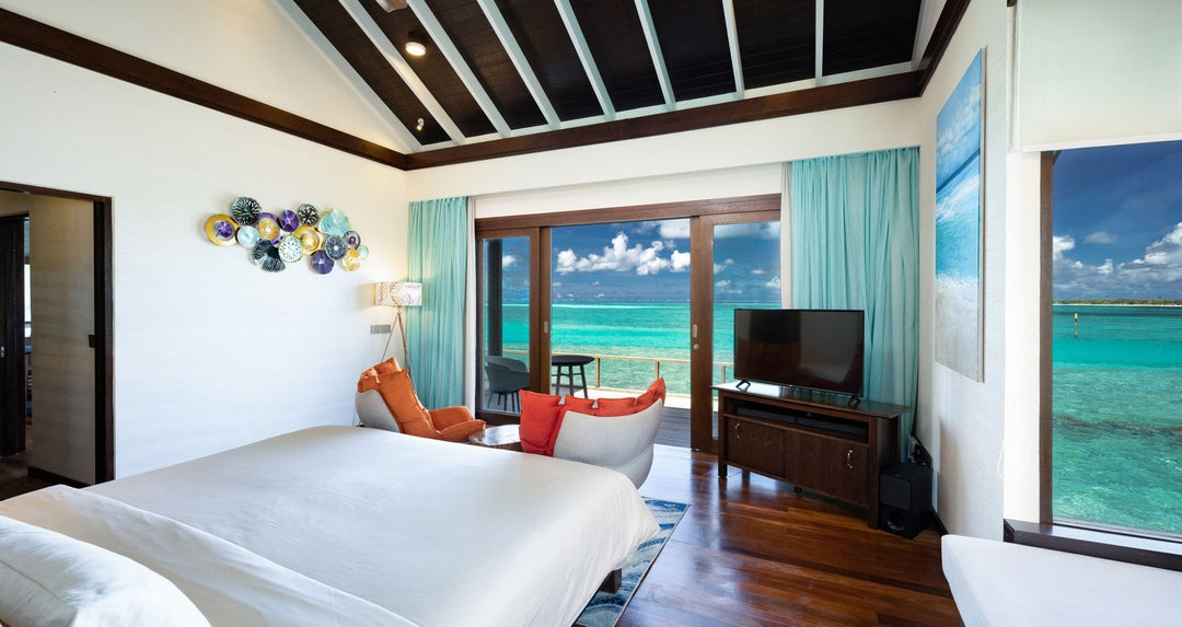 Atmosphere Kanifushi, Lhaviyani Atoll — an overwater villa seen from the bed, sliding doors folded back on to the deck rail and open water beyond.