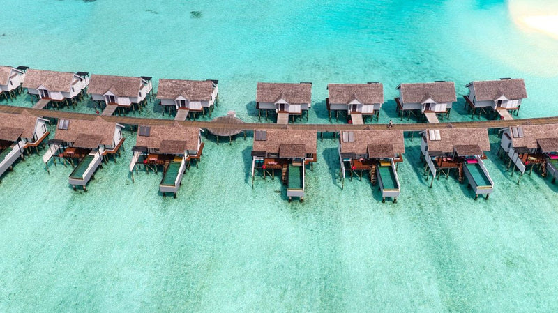 The overwater villas at OZEN LIFE Maadhoo from the air, thatched roofs along the jetty over pale turquoise water