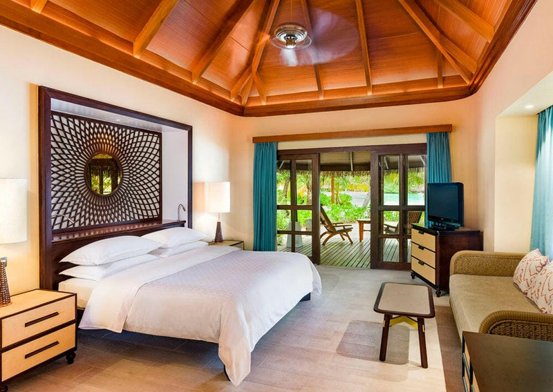A Beach Front Cottage bedroom at Sheraton Maldives Full Moon, a carved headboard and round mirror under timber.