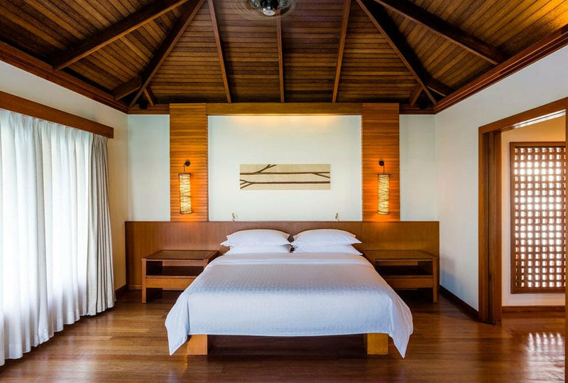 A water villa bedroom at Sheraton Maldives Full Moon, a dark timber headboard panel lit by wall lamps.