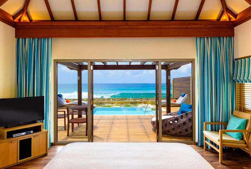 A villa living area at Sheraton Maldives Full Moon, teal curtains framing doors onto a lounger terrace.