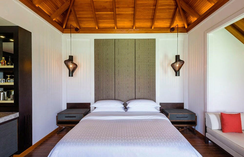 A water villa bedroom at Sheraton Maldives Full Moon, a tall padded headboard between two wall sconces.