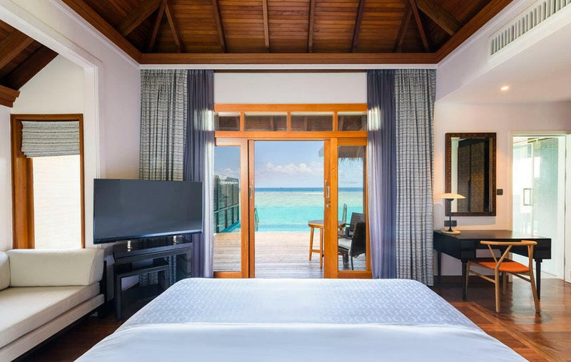 A water villa interior at Sheraton Maldives Full Moon, timber doors framing the turquoise lagoon past the bed.