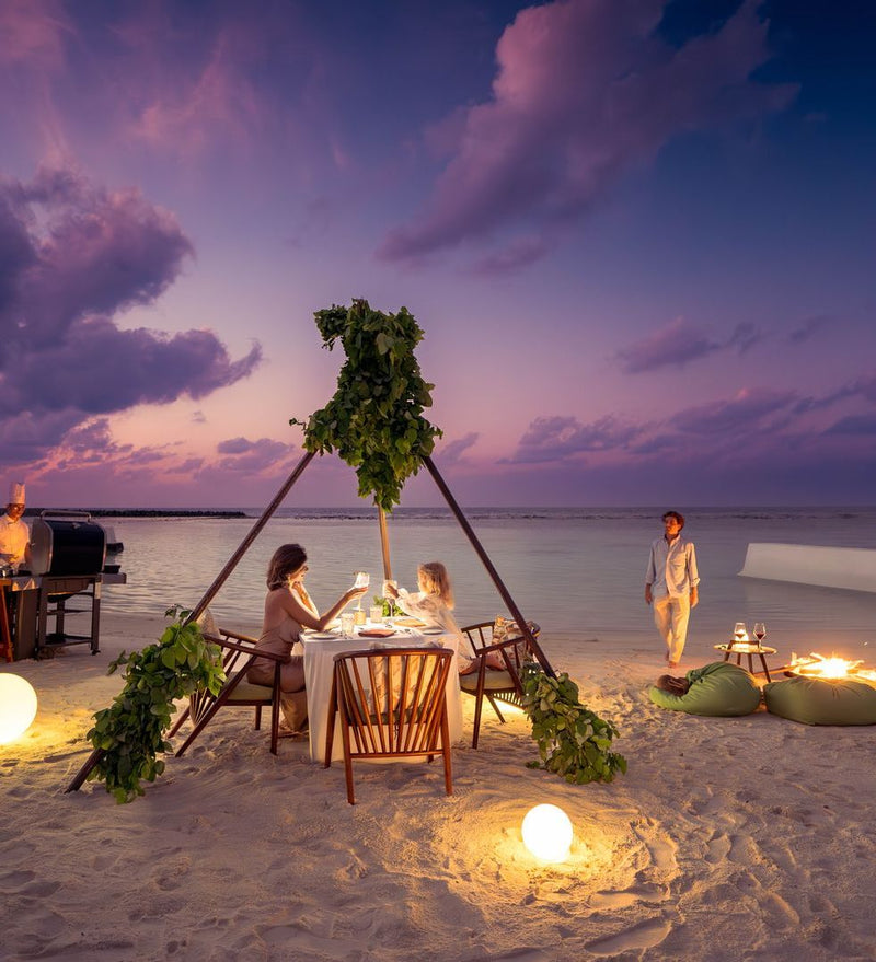 OBLU SELECT Sangeli, North Malé Atoll: a family dinner on the beach at dusk, the table set under a palm-frond frame