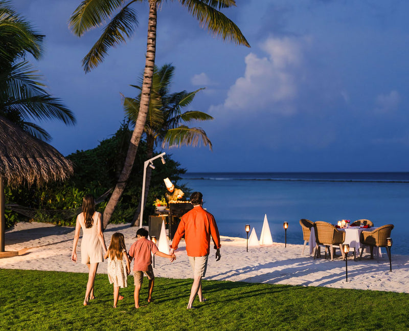 RAAYA by Atmosphere, Raa Atoll: a family walking to a candlelit dinner table on the beach at dusk