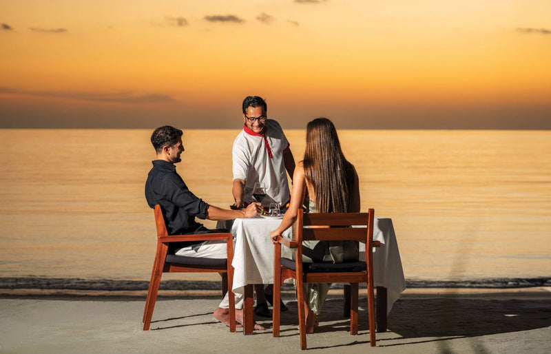 VARU by Atmosphere, North Malé Atoll: a waiter serving a couple at a beach table at sunset