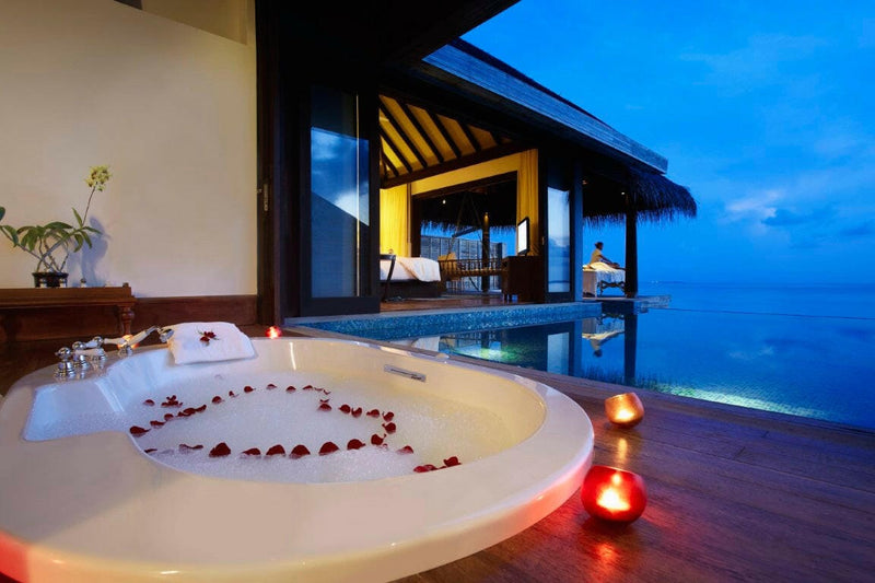 An Overwater Pool Villa bathroom at Anantara Kihavah Maldives Villas in the evening, the bath dressed with rose petals