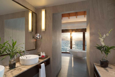 An Over Water Suite bathroom at Anantara Dhigu, twin basins and a bath looking out to the sea at sunset