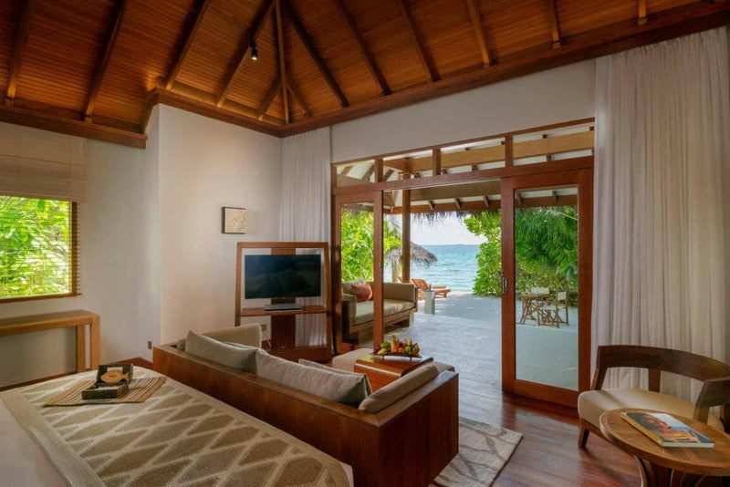 A beach villa living area at Baros Maldives, a low sofa under dark rafters facing the open beach doors.