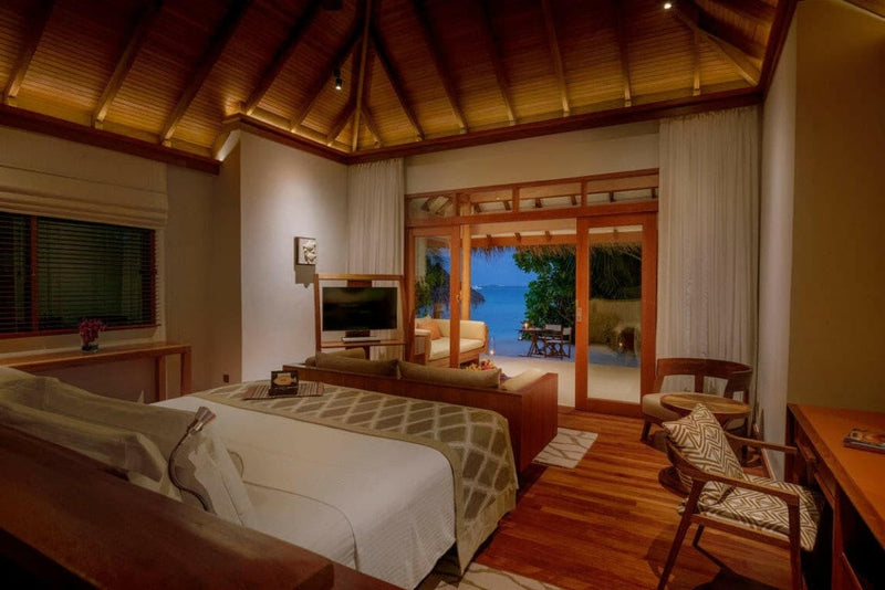 A beach villa bedroom at Baros Maldives, a striped runner on the bed with the sea through wide doors.