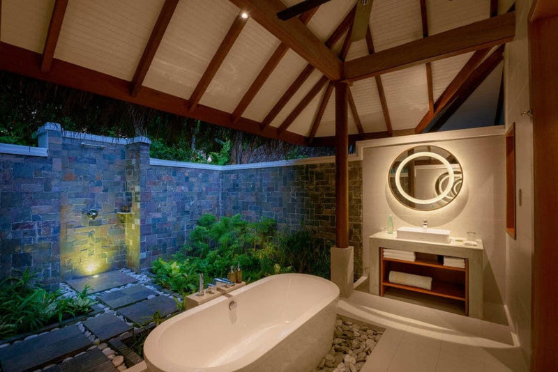An open air villa bathroom at Baros Maldives, an oval tub against a dark stone wall hung with planting.