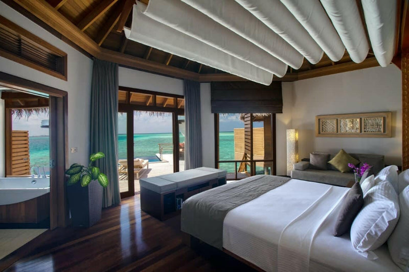 A water villa bedroom at Baros Maldives, blue cushions and a daybed facing the lagoon through glass.