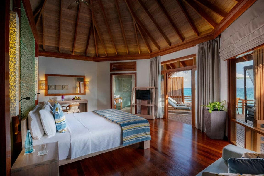A water villa bedroom at Baros Maldives, blue cushions and a striped throw on dark timber floors.