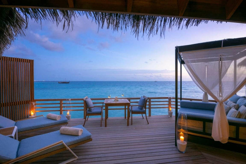 A water villa deck at Baros Maldives at dusk, a curtained daybed and loungers above a flat calm sea.