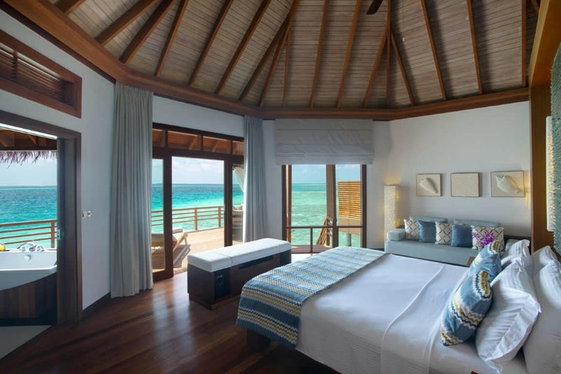 A water villa bedroom at Baros Maldives, doors folded back to the deck and the turquoise lagoon.