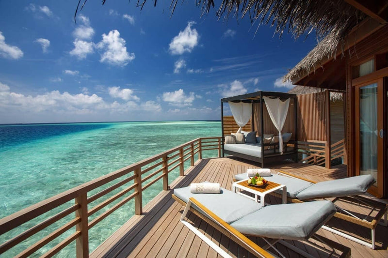 A water villa deck at Baros Maldives, two loungers and a curtained daybed above bright turquoise water.