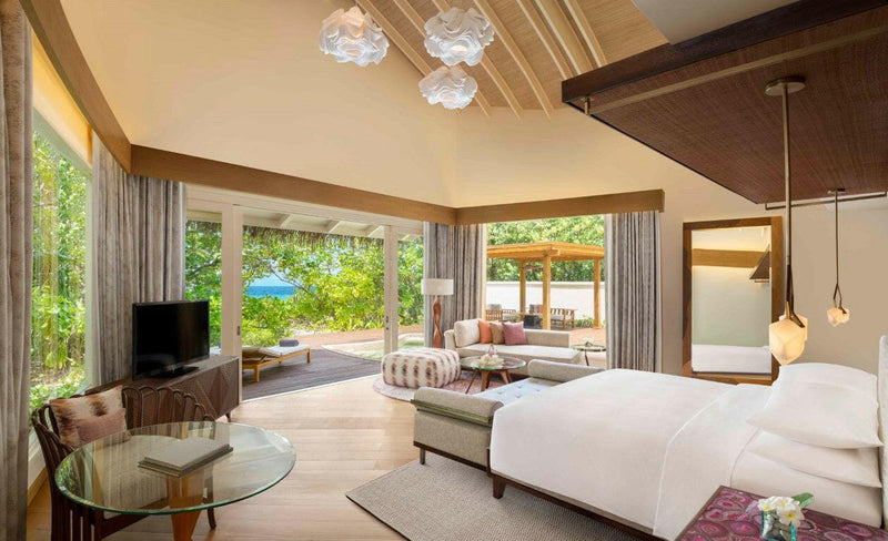 A beach villa bedroom at JW Marriott Maldives, pink cushions and a shaded pavilion beyond the doors.