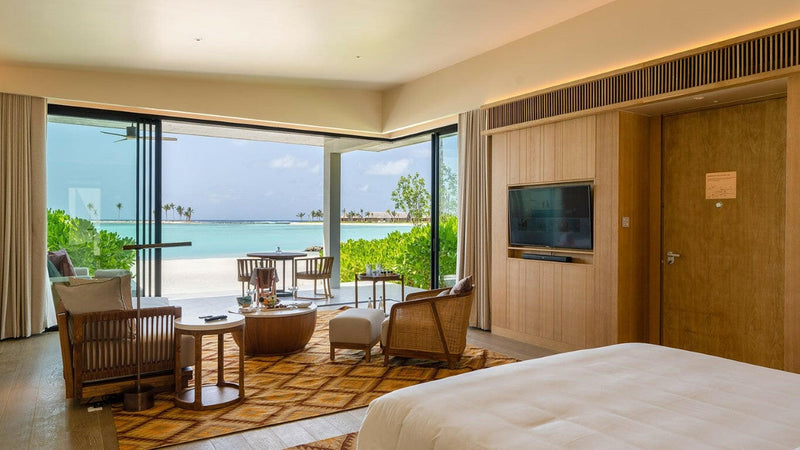 A beach villa room at Kuda Villingili, armchairs by glass doors onto a terrace and white sand.