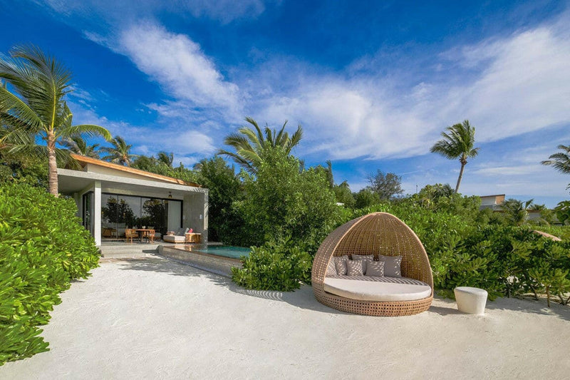 A round rattan daybed on the sand at Kuda Villingili, villa and pool behind under a cloudy sky.