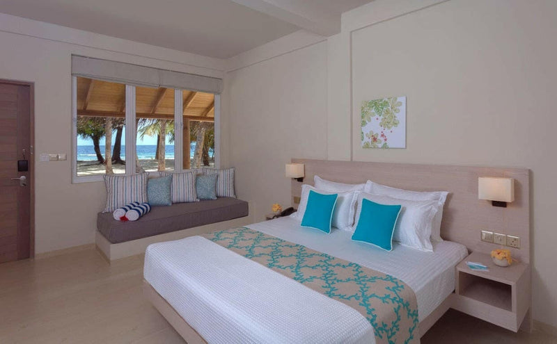 A Deluxe Room at Malahini Kuda Bandos with a king bed and a cushioned bench under windows onto the beach.