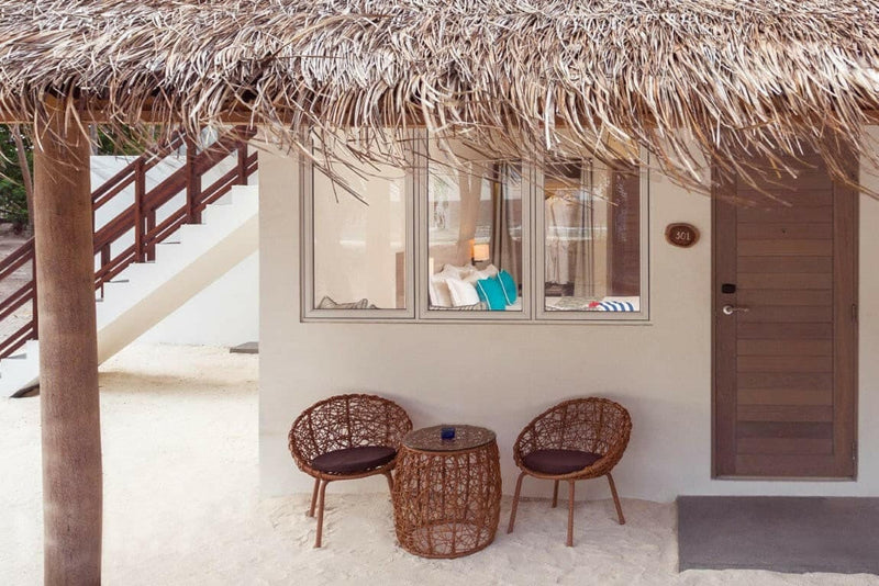 A thatched-roof room exterior at Malahini Kuda Bandos with its entrance door and two woven chairs on the sand.