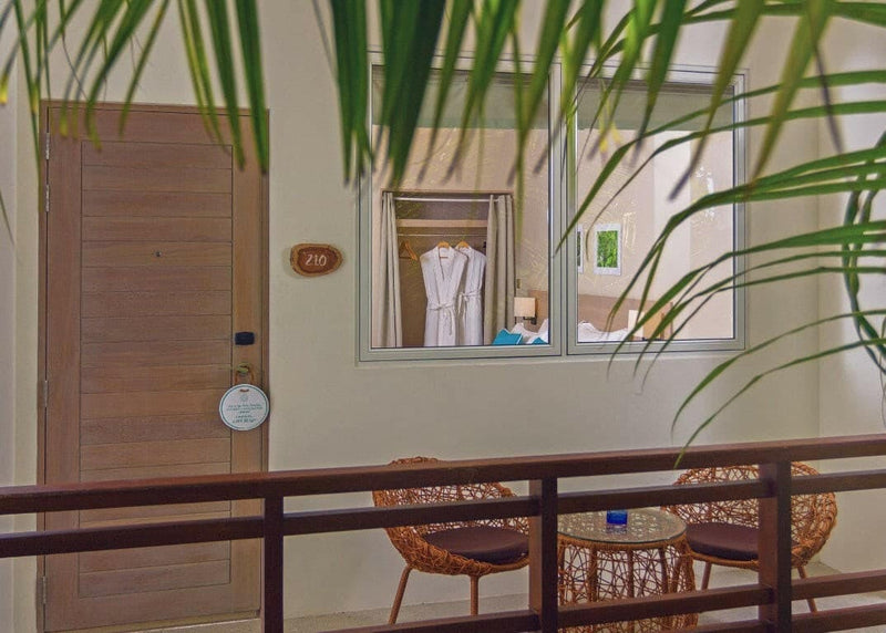 A room entrance at Malahini Kuda Bandos with its number plate, seen past two woven chairs and a table on a decked terrace.