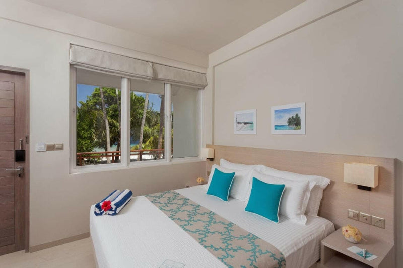 A Superior Room at Malahini Kuda Bandos with a king bed beside a window looking out to trees and the lagoon.