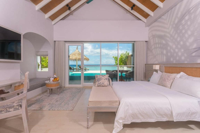 A suite bedroom at Malahini Kuda Bandos under a high vaulted ceiling, with sliding doors onto a terrace and the lagoon.